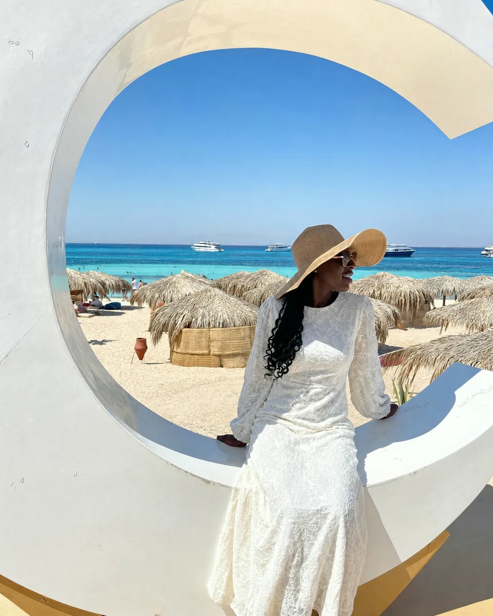 Precious Mensah, founder of Selah Journeys, in a white lace dress and a wide sun hat, sitting in a large white sculpture on a sunny beach